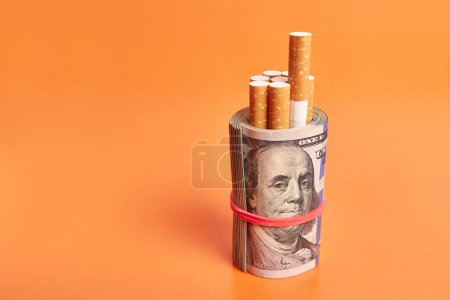 Photo for Cigarettes wrapped in 100 dollar bills on an orange background. Concept of expensive cigarettes. - Royalty Free Image