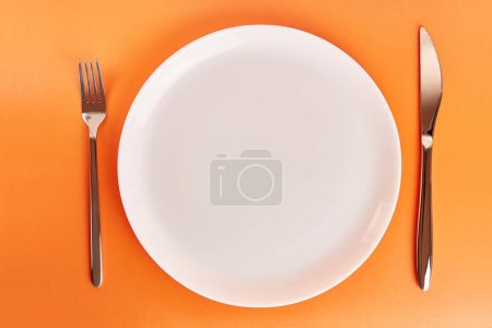 Téléchargez les photos : Assiette blanche avec fourchette et couteau sur fond orange. - en image libre de droit