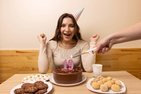 A girl sits in front of a table with a festive cake, in which a candle is lit in the form of the number 19. The concept of a birthday celebration.