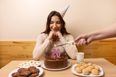 Foto de Una niña se sienta frente a una mesa con un pastel festivo, en el que se enciende una vela en forma de número 20. El concepto de una celebración de cumpleaños. - Imagen libre de derechos