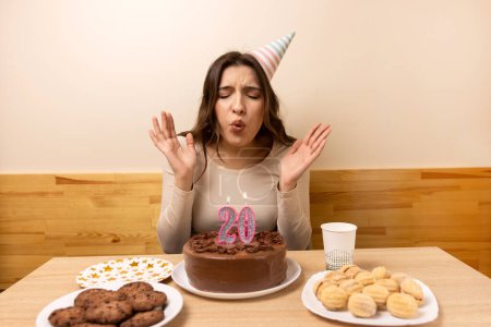 Foto de Una chica se sienta frente a una mesa con un pastel festivo, en el que sopla una vela en la forma del número 20. El concepto de una celebración de cumpleaños. - Imagen libre de derechos