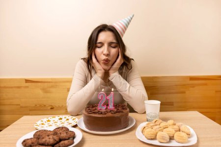 A girl sits in front of a table with a festive cake, in which she blows out a candle in the form of the number 21. The concept of a birthday celebration.