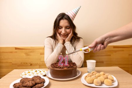 Foto de Una niña está sentada frente a una mesa con un pastel festivo, en el que se enciende una vela en forma de número 21. El concepto de una celebración de cumpleaños. - Imagen libre de derechos