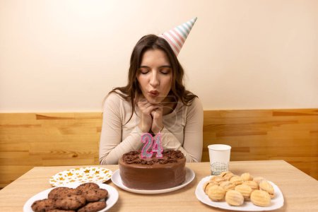 Foto de Una chica se sienta frente a una mesa con un pastel festivo y sopla una vela en forma de número 24. El concepto de una celebración de cumpleaños. - Imagen libre de derechos