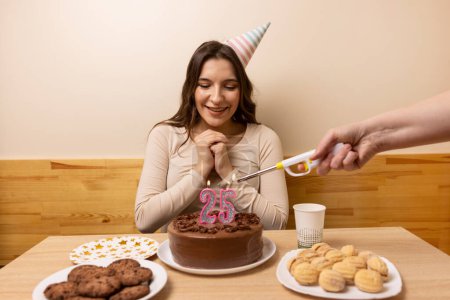 A girl is sitting in front of a table with a festive cake, on which a candle is burning in the form of the number 25. The concept of a birthday celebration.