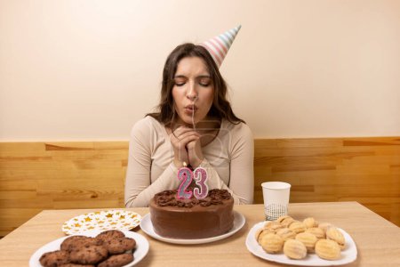 Foto de Una chica se sienta frente a una mesa con un pastel festivo y sopla una vela en forma de número 23. El concepto de una celebración de cumpleaños. - Imagen libre de derechos