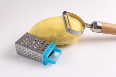 Peeled raw potatoes, peeling knife and grater on a white background.
