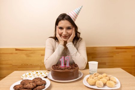 Photo for A girl sits in front of a table with a festive cake, in which a candle in the form of the number 17 has blown out. The concept of a birthday celebration. - Royalty Free Image