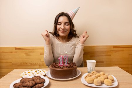 Photo for A girl sits in front of a table with a festive cake, in which a candle is lit in the form of the number 17. The concept of a birthday celebration. - Royalty Free Image