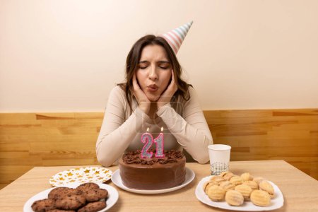 Foto de Una chica se sienta frente a una mesa con un pastel festivo, en el que sopla una vela en la forma del número 21. El concepto de una celebración de cumpleaños. - Imagen libre de derechos