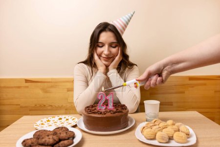 A girl is sitting in front of a table with a festive cake, in which a candle is lit in the form of the number 21. The concept of a birthday celebration.