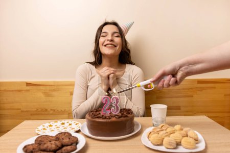 A girl sits in front of a table with a festive cake and a burning candle in the form of the number 23. The concept of a birthday celebration.