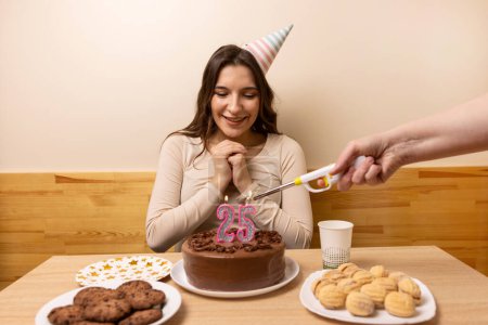 A girl is sitting in front of a table with a festive cake, on which a candle is burning in the form of the number 25. The concept of a birthday celebration.