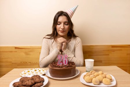 Foto de Una chica se sienta frente a una mesa con un pastel festivo, en el que sopla una vela en la forma del número 26. El concepto de una celebración de cumpleaños. - Imagen libre de derechos