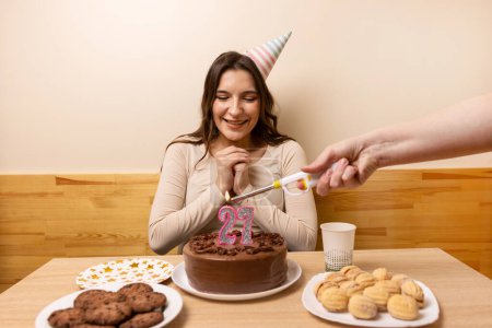 Foto de Una chica se sienta frente a una mesa con un pastel festivo, en el que sopla una vela en forma de número 27. El concepto de una celebración de cumpleaños. - Imagen libre de derechos