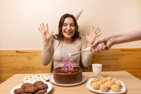 Téléchargez les photos : Une fille s'assied devant une table avec un gâteau festif, sur lequel une bougie est allumée sous la forme du numéro 26. Le concept d'une célébration d'anniversaire. - en image libre de droit