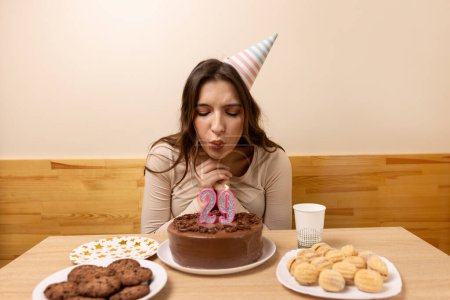A girl sits in front of a table with a festive cake, on which she blows out a candle in the form of the number 29. The concept of a birthday celebration.