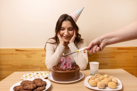 A girl sits in front of a table with a festive cake, on which a candle is lit in the form of the number 28. The concept of a birthday celebration.