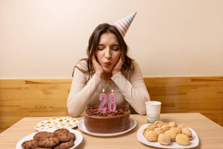 Foto de Una chica se sienta frente a una mesa con un pastel festivo, en el que sopla una vela en la forma del número 28. El concepto de una celebración de cumpleaños. - Imagen libre de derechos