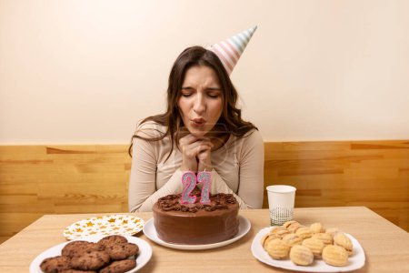 Foto de Una chica se sienta frente a una mesa con un pastel festivo, en el que sopla una vela en forma de número 27. El concepto de una celebración de cumpleaños. - Imagen libre de derechos