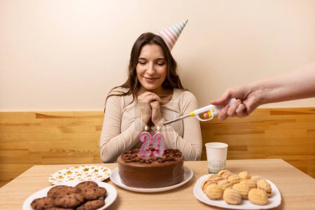 A girl is sitting in front of a table with a festive cake, on which a candle is lit in the form of the number 29. The concept of a birthday celebration.