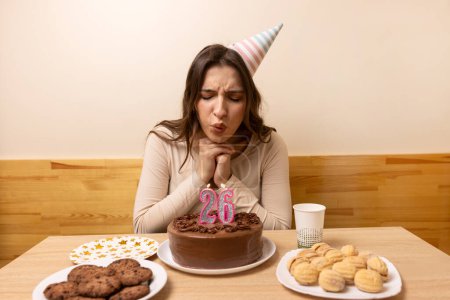 A girl sits in front of a table with a festive cake and blows out a candle in the form of the number 26. The concept of a birthday celebration.