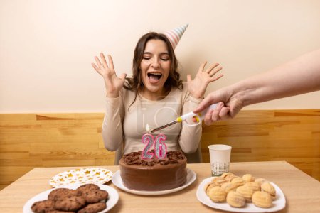 Foto de Una niña se sienta frente a una mesa con un pastel festivo, en el que se enciende una vela en forma de número 26. El concepto de una celebración de cumpleaños. - Imagen libre de derechos