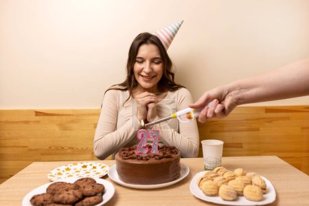 A girl sits in front of a table with a festive cake, on which she blows out a candle in the form of the number 27. The concept of a birthday celebration.
