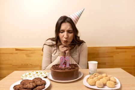 A girl sits in front of a table with a festive cake and blows out a candle in the form of the number 27. The concept of a birthday celebration.