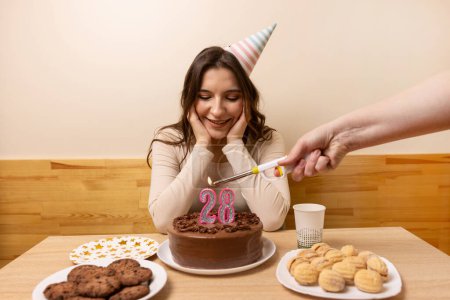 Foto de Una niña se sienta frente a una mesa con un pastel festivo, en el que se enciende una vela en forma de número 28. El concepto de una celebración de cumpleaños. - Imagen libre de derechos