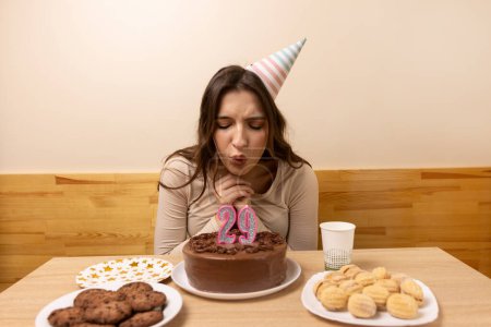 A girl sits in front of a table with a festive cake on which she blows out a candle in the form of the number 29. The concept of a birthday celebration.