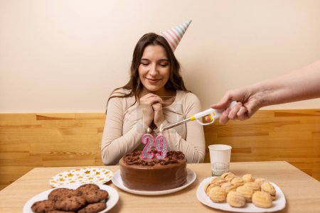 A girl sits in front of a table with a festive cake on which a candle is lit in the form of the number 29. The concept of a birthday celebration.