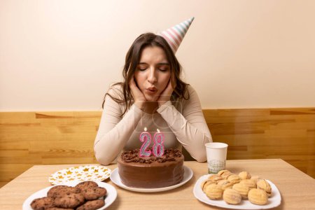 A girl sits in front of a table with a festive cake and blows out a candle in the form of the number 28. The concept of a birthday celebration.