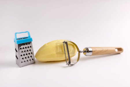 Peeled raw potatoes, peeling knife and grater on a white background.