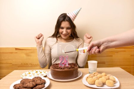 A girl is sitting in front of a table with a festive cake, in which a candle is lit in the form of the number 24. The concept of a birthday celebration.