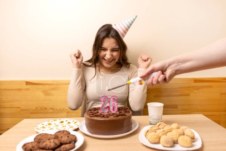 Ein Mädchen sitzt vor einem Tisch mit einer festlichen Torte, auf der eine Kerze in Form der Zahl 26 entzündet wird. Das Konzept einer Geburtstagsfeier.