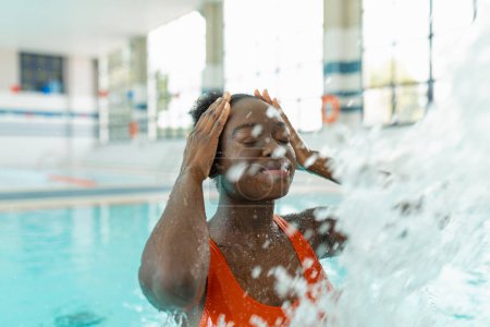 Foto de Mujer joven negra tocándose la cabeza mientras se relaja bajo el chorro de agua en la piscina climatizada durante un día de invierno - Imagen libre de derechos