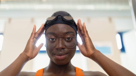 Foto de Mujer joven negra que se pone las gafas de baño sentada en el vestuario de la piscina climatizada - Imagen libre de derechos