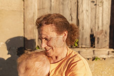 Photo for A young grandmother holds her baby granddaughter, just a few months old, in her arms during a sunny summer afternoon in a small town in the north of Galicia, Spain - Royalty Free Image