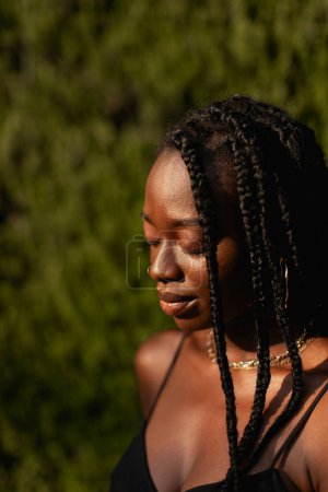 Foto de Retrato de una joven negra con trenzas en el pelo mientras disfruta de la tarde relajándose con los ojos cerrados, fondo verde borroso - Imagen libre de derechos