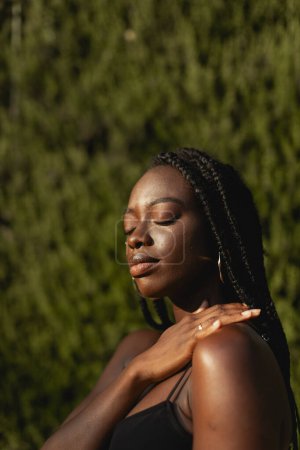 Foto de Una joven negra con trenzas en el pelo, tocándose el hombro con la mano, los ojos cerrados, fondo verde borroso - Imagen libre de derechos