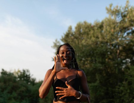 Foto de Una joven negra con trenzas en el pelo ondea y saluda durante un paseo por el parque al atardecer - Imagen libre de derechos