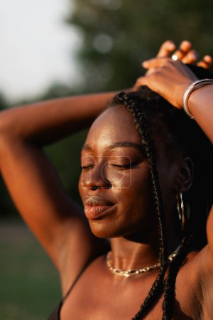 Téléchargez les photos : Une jeune femme noire avec des tresses, touche ses cheveux, se sent belle, et profite du soleil lors d'une promenade au coucher du soleil - en image libre de droit