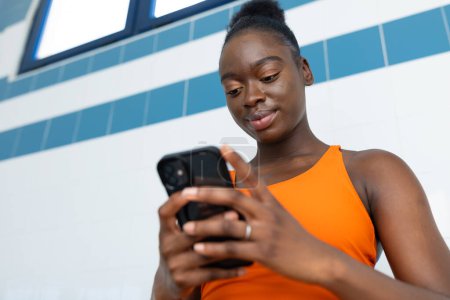 Photo for Black young woman holding her smartphone with both hands while sitting in a bench at the heated swimming pool - Royalty Free Image
