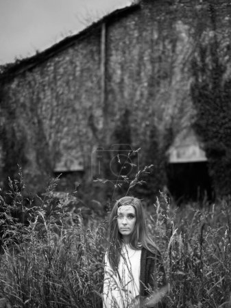 Foto de Una joven mujer mira a la cámara, en un lugar salvaje, escondido entre hierbas altas, cerca de un lugar abandonado - Imagen libre de derechos