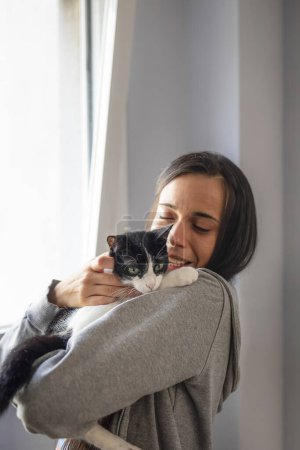 Téléchargez les photos : Une jeune femme s'étreint et joue avec son chat noir et blanc à la maison. - en image libre de droit