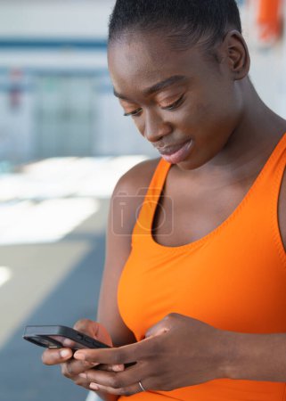 Photo for Black young woman using her smartphone, sitting in a bench at the heated swimming pool - Royalty Free Image
