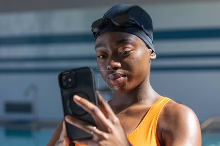 Photo for Black young woman holding her smartphone while standing at the heated swimming pool - Royalty Free Image