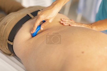 Foto de Una fisioterapeuta femenina masajea la espalda de un hombre mayor con una herramienta de masaje, un protector de dedos, durante una sesión de terapia manual en una clínica - Imagen libre de derechos