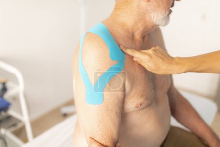 Foto de Una fisioterapeuta hembra saca el vendaje neuromuscular o cinta de cinesiología del hombro de su paciente, un hombre mayor - Imagen libre de derechos